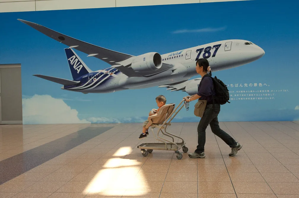 daytime landscape view of a woman and a child in a stroller walking in front of an 'ana 787' advertisement at the haneda airport in tokyo